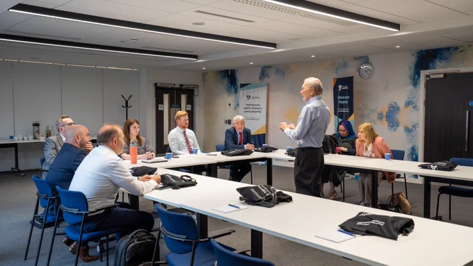 Eight people sit around table benches as a man wearing a light shirt and dark trousers stands in the middle leading a workshop session.