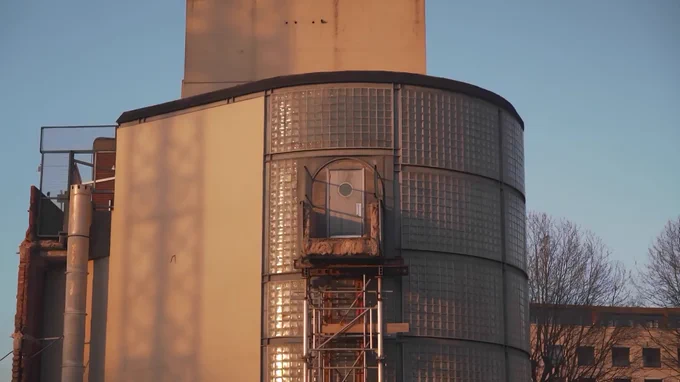 Industrial silver cylindrical tower with a door in the middle, possibly a gas holder or a water tower. There is a building in the background and trees without leaves.