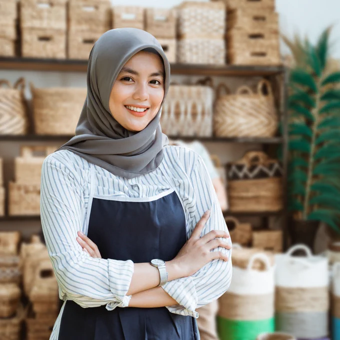 Person wearing a hijab and apron standing with arms folded inside a retail shop selling woven baskets and homeware items.