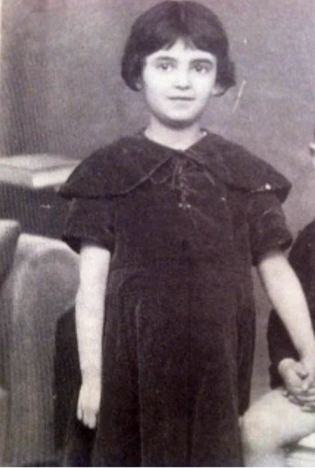 A vintage black and white photograph of a young girl standing, dressed in a dark collared dress with a ruffled neckline, looking directly at the camera. The background is slightly blurred, emphasizing the subject in the foreground.
