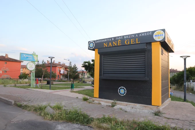 Fotoğrafta bir parkın girişine yakın konumda bulunan küçük bir satış veya hizmet büfesi görülüyor. Büfe siyah ve sarı renklerde tasarlanmış; ön cephesindeki kepenk kapalı durumda. Üst bölümünde Kürtçe “Nanê Gel” yazısı ile çeşitli kurum logoları yer alıyor.

Sol tarafta “Mimoza Parkı” yazılı bir yönlendirme tabelası bulunuyor. Büfenin çevresinde yaya yolları, çim alanlar, aydınlatma direkleri ve ağaçlar görülüyor. Arka planda iki ve üç katlı konut binaları yer alıyor. Açık havada çekilen fotoğrafta gökyüzü açık ve aydınlık. Yol kenarında konumlanan yapının çevresinde az sayıda kişi uzaktan seçilebiliyor. Fotoğraf, park çevresindeki kentsel bir kamusal alanı gösteriyor.