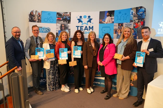 A group of nine individuals stand in front of a backdrop displaying the "TEAM MODE SHIFT STARS" logo and images of people using various modes of transportation. The individuals, consisting of five women and four men, are holding certificates and wooden awards, indicating they are award recipients. The backdrop also features the text "MODE SHIFT MONTH" multiple times, suggesting an event or recognition related to sustainable transportation.