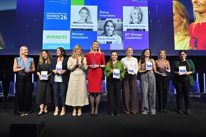 An onstage photograph of nine women standing in a line at the European Prize for Women Innovators 2026. A large screen behind them displays the event title and the names and portraits of category winners: Katerina Spranger (Women Innovators), Marta Oliveira (Rising Innovators), and Ella Frances Cullen (EIT Women Leadership). The women on stage are holding awards and booklets, smiling and celebrating.
