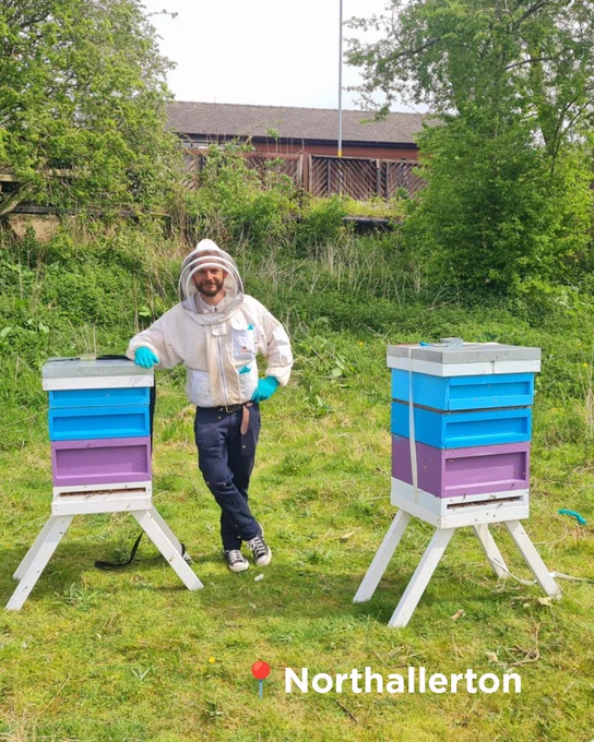 A beekeeper in a white suit stands next to a colourful beehive in a grassy field.