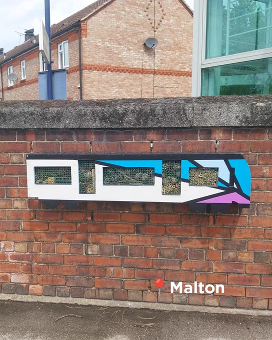 An insect hotel in the shape of a class 185 train with a colourful geometric design mounted on a brick wall.