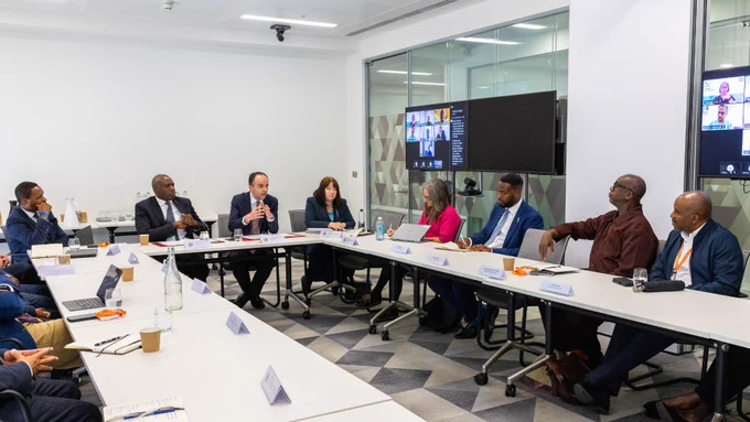 A group of people are seated around a U-shaped conference table in a modern meeting room. A screen displays a video call with additional participants. 
