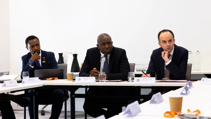 A group of individuals are seated at a conference table engaged in discussion. Papers, pens, and drinks are on the table. The setting appears formal and professional.