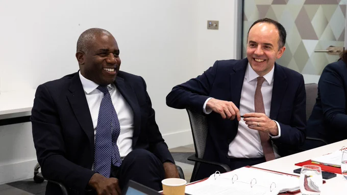 Two individuals are seated at a table in an office setting, engaged in a meeting. They are both smiling. Papers, a paper cup, and a glass of water are visible on the table. 