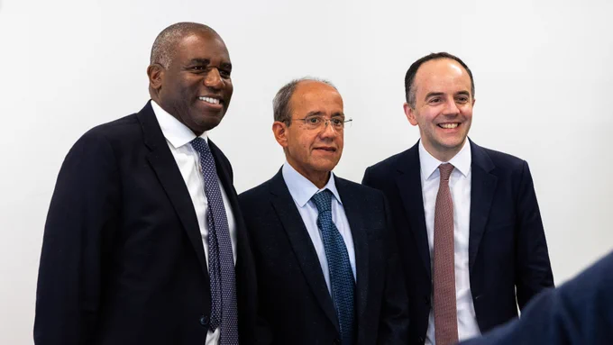 Three individuals in business attire are standing together against a plain background, all smiling.