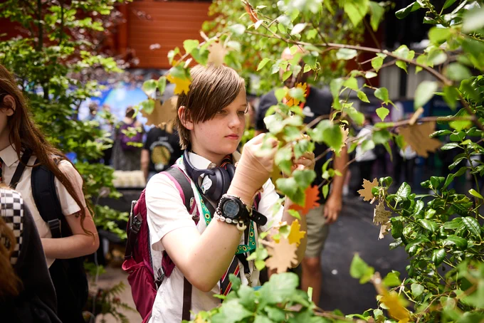 A child in school uniform examines leaves on a bush. They're wearing a backpack, headphones around their neck, and a lanyard with BBC Earth branding. The background shows more foliage and some blurred individuals.