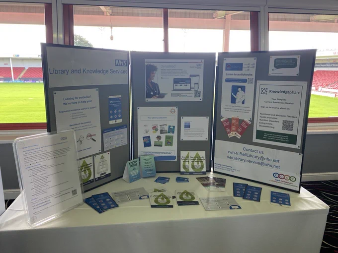 NHS Library and Knowledge Services promotional display booth at an indoor event. A three-panel grey display board stands behind a white-clothed table, featuring NHS branding, posters for DynaMed, evidence search services, audiobooks and KnowledgeShare resources. Various pamphlets, flyers, and informational cards with green droplet logos are arranged on the table. In the background, large windows reveal a green sports stadium field with red seating.