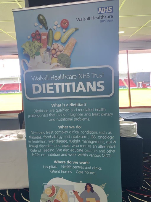 NHS Walsall Healthcare NHS Trust pull-up banner promoting Dietitians. The blue and teal banner features images of fresh healthy foods (avocado, vegetables, fruit, eggs, bread) at the top, followed by the heading “DIETITIANS”. It explains what dietitians do, the clinical conditions they treat (diabetes, IBS, oncology, malnutrition, etc.), and where they work (hospitals, clinics, patient homes, care homes). An illustration of a smiling dietitian appears at the bottom. The banner is displayed indoors with a stadium visible through the windows in the background.