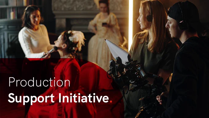 Film crew members collaborate on set, preparing equipment and touching up an actors make-up on the set of a period drama. In the lower left is a semi-opaque, curved red overlay with the words 'Production Support Initiative' in white.