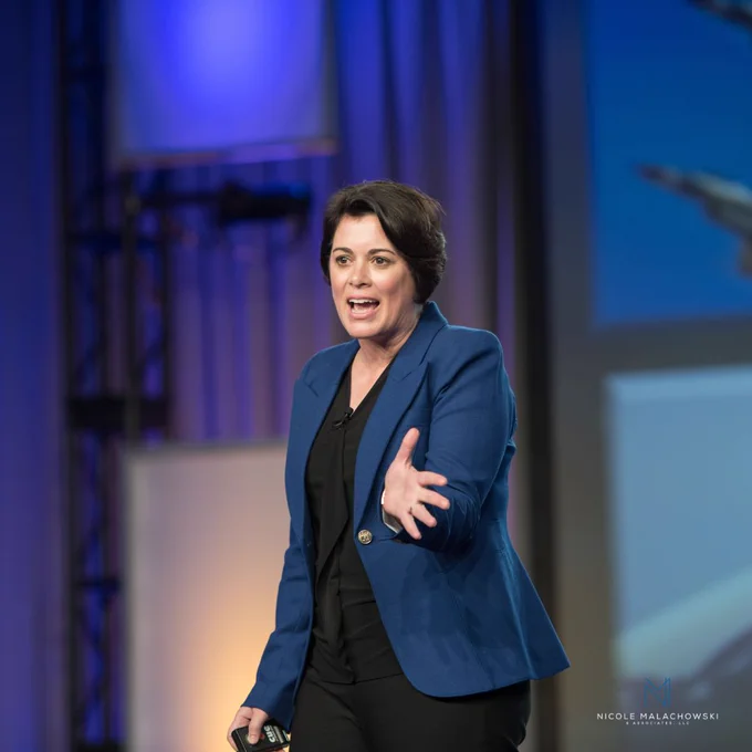 Nicole Malachowski is delivering a keynote-style presentation on stage, gesturing toward the audience with an expressive, engaged posture. She’s wearing a bright blue blazer over a black outfit and appears to be speaking passionately into a headset or lapel microphone. The background is softly lit with blue and purple stage lighting, creating a professional conference atmosphere. A presentation screen is partially visible behind her, and the image includes a “Nicole Malachowski & Associates, LLC” watermark in the bottom-right corner.
