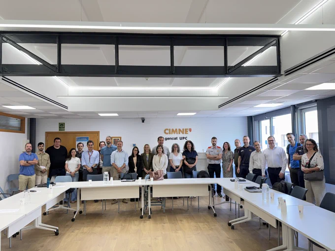 Un grup d'unes vint persones de peu juntes en una sala de reunions lluminosa a la seu de CIMNE a Barcelona. Estan situades darrere d'un conjunt de taules blanques disposades en forma de U, somrient i mirant cap a la càmera. La paret del darrere mostra el logotip de CIMNE juntament amb "gencat UPC", que indica la col·laboració entre el Centre Internacional de Mètodes Numèrics en Enginyeria i el Govern de la Generalitat de Catalunya. Els assistents inclouen representants de CIMNE i del Departament d'Interior i Seguretat Pública de la Generalitat de Catalunya, alguns dels quals porten uniformes de protecció civil i serveis d'emergències. La sala moderna, amb sòl de fusta i grans finestres, transmet una atmosfera professional i col·legial després d'una sessió de treball productiva sobre gestió d'emergències i protecció civil.