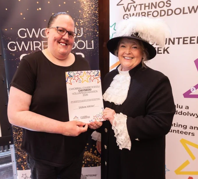 Two people smiling holding a piece of paper that reads 'Gwobrau Gwirfoddoli Gwynedd Volunteer Awards 2026'