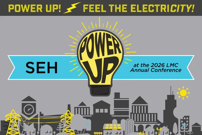 Yellow light bulb with 'POWER UP' text glowing above a cityscape, promoting the 2026 LMC Annual Conference by SEH.