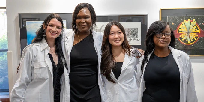 Four graduates standing side by side indoors, wearing white coats over black dresses, posing in front of framed artwork.