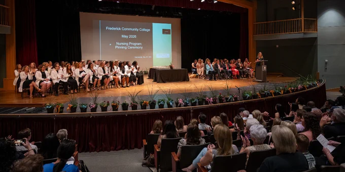 Graduates in white coats seated on stage during Frederick Community College Nursing Program Pinning Ceremony with audience watching.