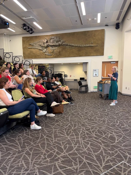 A blonde female presenter speaking to an audience seated in a tiered lecture hall with green seats. She stands at the front of the room next to a gray utility cart, gesturing as she speaks. Positioned high on the wall behind the audience is a large, complete fossil skeleton of a prehistoric whale.