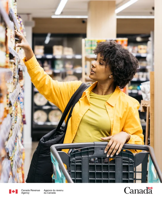 A person shopping for groceries reaches for an item on the shelf while pushing a shopping cart.