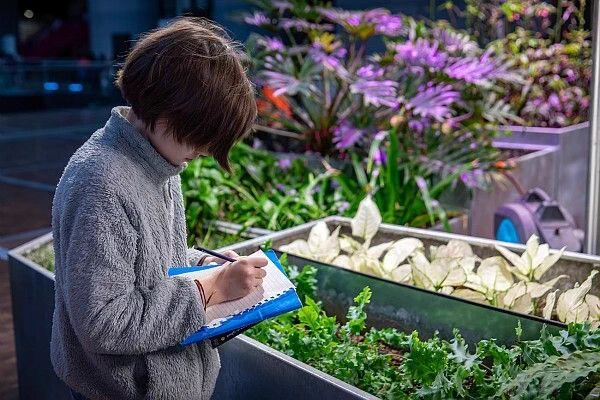 Enfant prenant des notes dans un carnet devant des bacs remplis de plantes et de végétation à la Cité des sciences et de l’industrie, dans une ambiance lumineuse et verdoyante propice à l’observation du vivant.