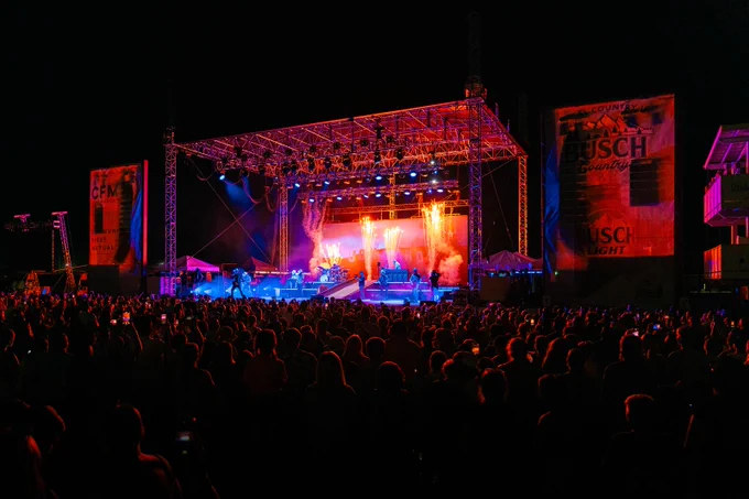 A large crowd watches a live concert on an outdoor stage at night, with colorful lights illuminating the performers and stage banners visible on both sides.
