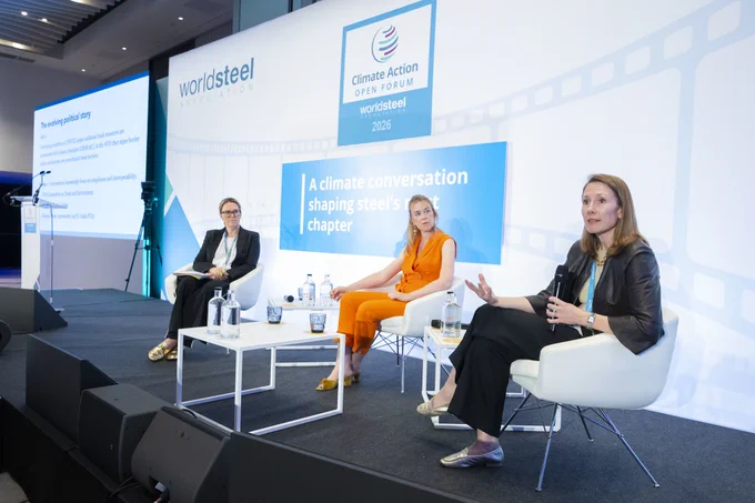 Three women sit on a stage during a panel. One woman holds a microphone and speaks to an audience (not pictured). The other two women watch her as she speaks and listen.
