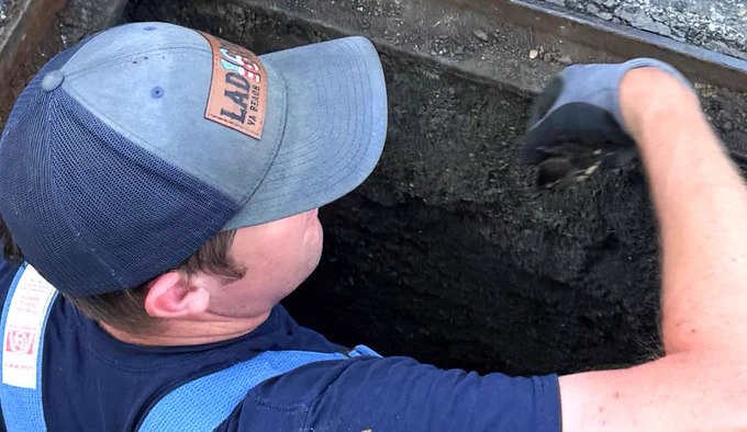 An individual wearing a cap is inspecting or working inside a large, rectangular pit. They are wearing gloves and a dark shirt.