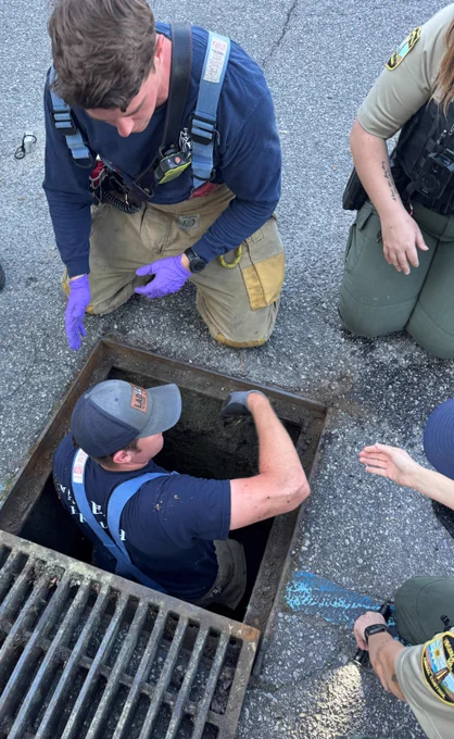 A person wearing a uniform, partially inside a manhole, is being assisted by others. The setting appears to be a street area. There are visible badges and equipment, including gloves, indicating an emergency or rescue scenario.