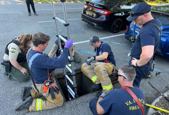 A group of people, including individuals in Virginia Beach Fire Department attire, are gathered around an open manhole in a parking lot. One person is using a ladder to descend into the manhole, while others observe and assist. Nearby are parked cars and a uniformed law enforcement official.