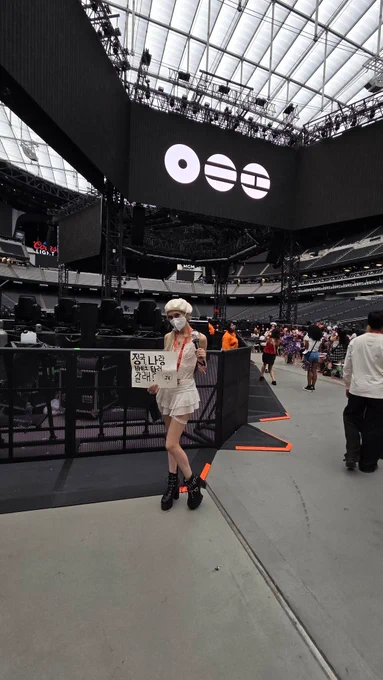 April masks, a mixed young woman in a white N95 mask, lace skirt, body suit, beret, and black boots standing in front of a stage holding a sign in Korean for JK at a BTS concert in Las Vegas.