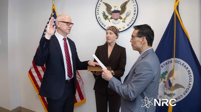 A man in a suit raises his right hand while taking an oath before two officials in front of U.S. Nuclear Regulatory Commission flags and seal.