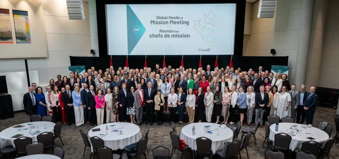 De nombreuses personnes dans une grande salle de conférence posant pour une photo de groupe devant un écran affichant ce texte : «Global Heads of Mission Meeting - Réunion des chefs de mission ».