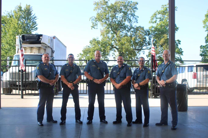 Six highway patrol men are smiling while looking at camera. The background contains trees and vehicles. 
