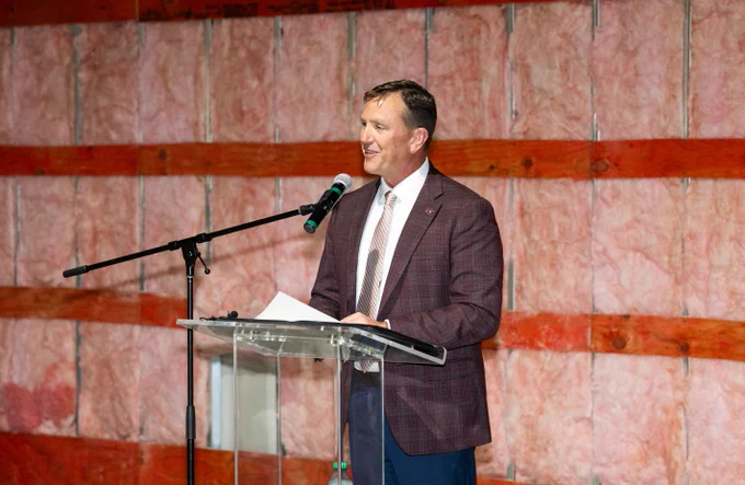 In the image, a person is speaking at a podium with a microphone. The backdrop has visible insulation and wooden beams.