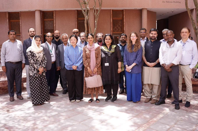 A group of people standing outdoors in front of The Habib Bank Building. They are arranged in two rows, with some smiling. The setting is sunny and shaded by trees.