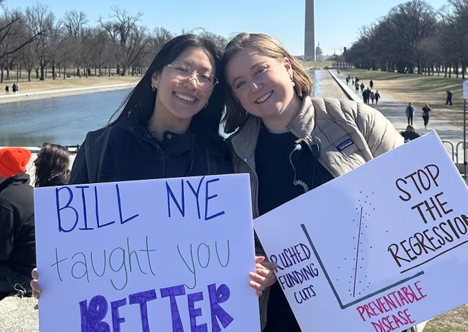 Two women smile and pose for a photo while holding protest signs about science in front of the reflecting pool in Washington, DC with the Washington Monument in the background. One sign says, "Bill Nye taught you better," and the other shows a graph that says, "Rushed funding cuts" on the y axis, "Preventable deaths" on the x axis, and "Stop the regression" next to the graph. 