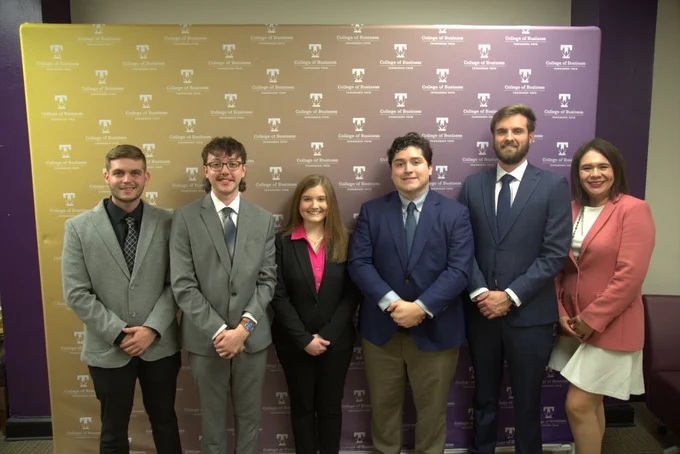 A group of six people wearing formal attire stands in front of a backdrop that reads "College of Business." The backdrop features a purple and yellow gradient with a logo.
