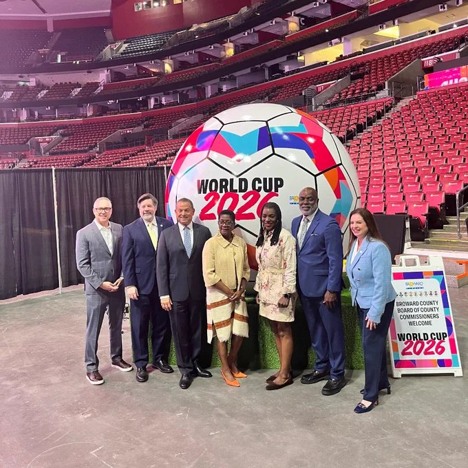 Group of six diverse professionals posing in front of a large colorful World Cup 2026 soccer ball inside an empty stadium.