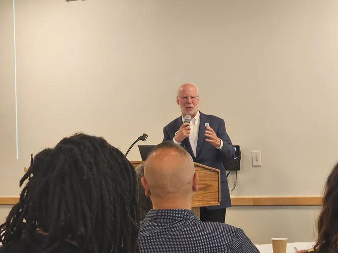 JB Holston speaks into a microphone from behind a wooden podium in a conference room.