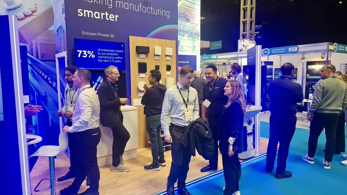 A group of people are gathered at a trade show booth featuring Ericsson's private 5G technology. The booth displays the slogan "making manufacturing smarter," alongside various 5G devices mounted on a wooden panel. A sign indicates that 73% of enterprises plan to include private 5G networks. Attendees are engaged in conversation and exploring the booth's exhibits.