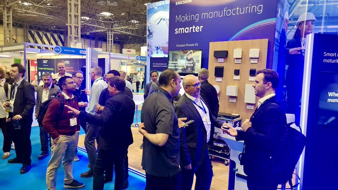 A busy exhibition booth at a trade show features a display for Ericsson, with the slogan "Making manufacturing smarter." Several people are engaged in conversation near the booth, which displays electronic devices. The setting is bright, with numerous other booths visible in the background.