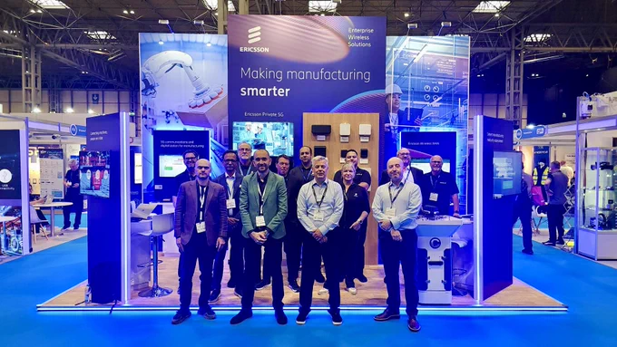 A group of people stand in front of the Ericsson booth at a trade show, with a display featuring technology and the slogan "Making manufacturing smarter." The setting is brightly lit, with various technological exhibits visible in the background.