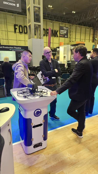 People are standing and interacting in an exhibition setting. One person is gesturing towards a display stand featuring inventory technology that uses drones. Ericsson branding is visible in the background. The environment suggests a professional or corporate event.