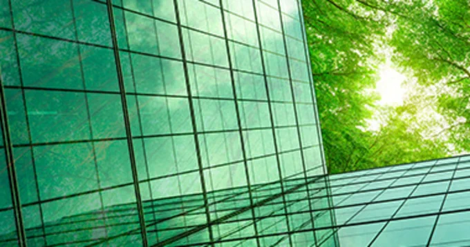 Modern glass building reflecting the bright green leaves of nearby trees on a sunny day.