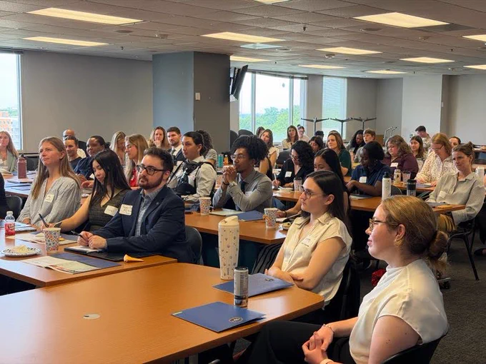 A diverse group of young adults sitting at rows of long wooden tables in a bright conference room or classroom during an orientation presentation. They are looking attentively toward the front left. In the foreground, a man with glasses and a navy blue blazer sits next to a woman with long dark hair. Pens, notebooks, folders, and water bottles are scattered across the desks.