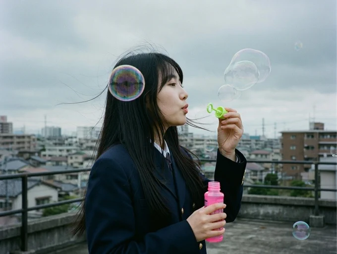 A girl in a dark school uniform stands outdoors on a rooftop, creating soap bubbles using a green bubble wand. She holds a pink bubble solution bottle, with large bubbles floating around her against a cloudy sky and city backdrop.