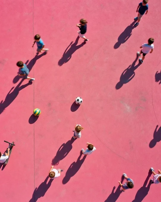 An aerial view of a pink playground with children playing. A group of kids is scattered across the surface, with some playing soccer near a soccer ball, while others engage with a colorful beach ball. Their shadows stretch across the ground.