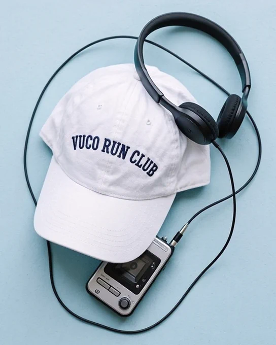 A white cap with 'VUCO RUN CLUB' embroidered in navy blue sits on a light blue surface. A pair of black headphones rests on the cap, with their cord connected to a silver portable cassette player below.
